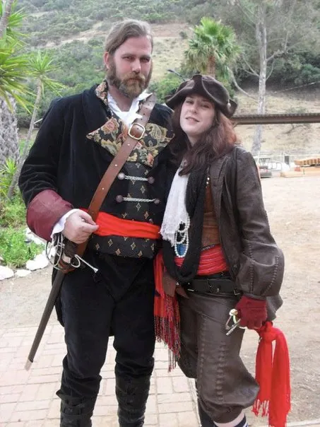 A couple in pirate costume, a bearded man in black velvet with a sword and a woman in a tricorn hat, leather jacket, and red sash