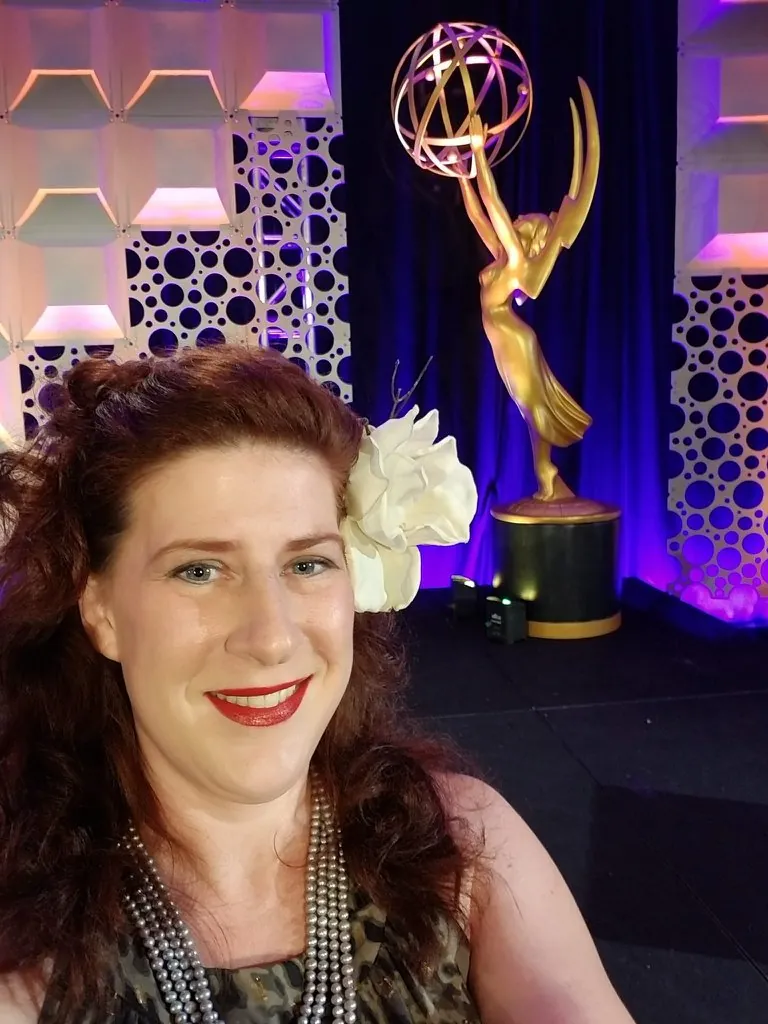 Mary Poplin smiling in front of a golden Emmy statue on a purple-lit stage