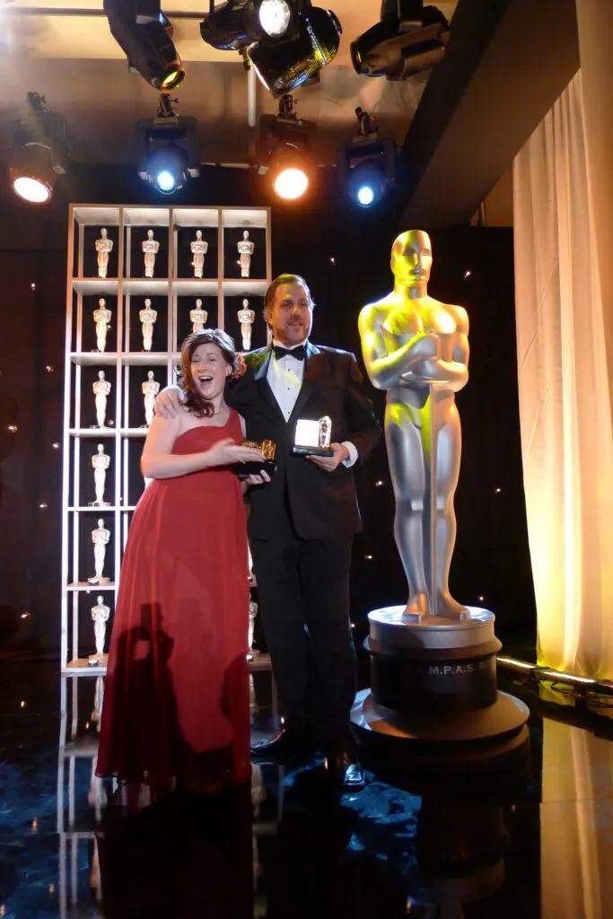 Mary Poplin and a colleague in formal wear beside a large Oscar statue and a shelf of statuettes